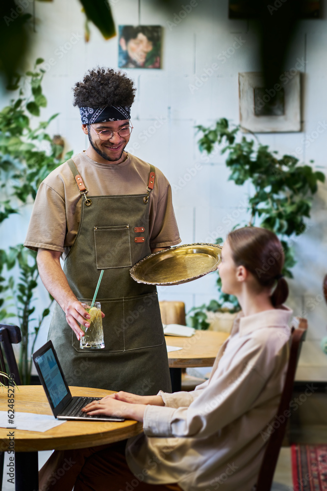 Poster young waiter in apron serving brunette female guest of cafe sitting by table and analyzing online da