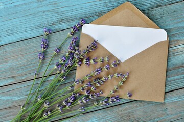 Mockup of craft paper envelope with blank white card with lavender flowers on blue old wooden background close-up. Happy Birthday, Valentine's day, wedding, Mother's Day greeting, invitation card.