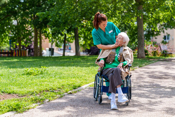 An elderly woman in a wheelchair is tenderly caressed by her caring nurse as they take a sunny...