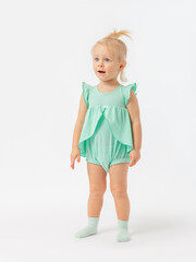 The child opened his mouth in surprise. A 2-year-old girl stands on a white background in socks and a green dress. Expressive look.