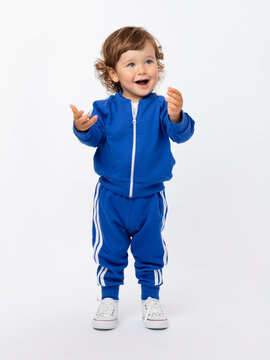 Joy, happiness, delight. A cute little boy of 1-2 years old with curly hair stands and emotionally moves his hands. Blue tracksuit and sneakers on a white background.