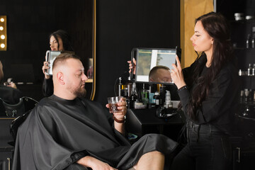 The master hairdresser shows the client a new hairstyle in the mirror. Man with a glass of drink