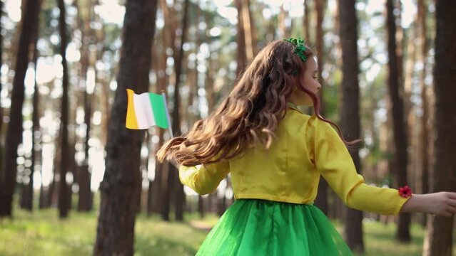 Cute little girl celebrating St Patricks Day at outdoor festival. Beautiful white child dressed as a fairy spinning around with Irish flags in hands