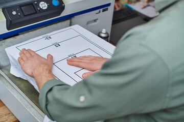Cropped view of young artisan working with t-shirt with marking and screen printing machine in print studio, customer-focused small business concept, graphic design