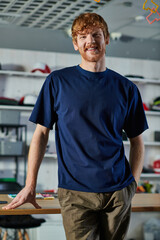 Portrait of cheerful redhead craftsman in casual clothes holding hand in pocket and smiling at camera while standing in print studio, hands-on entrepreneurship concept