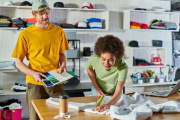 Smiling young african american craftswoman measuring clothes while working near colleague with clothes samples and laptop blurred in print studio, self-employment opportunity concept