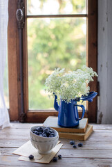 Vaso di latta con fiori di carota selvatica e tazza di mirtilli su un tavolo di legno di fronte ad una finestra