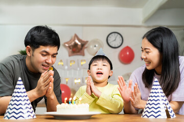 Happy birthday party in family, asian young parent father, mother and little cute boy or child celebrating, blowing candles on the cake and decorated balloons, celebration at home. Lovely moments