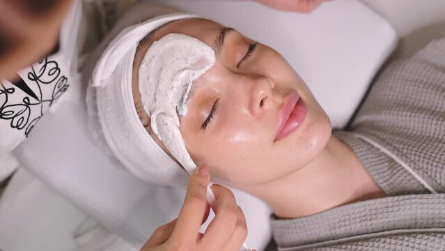 Video of a pretty woman lying in a salon, beautician applying moisturizing mask on her face. Moisturizing nourishing facial mask, scrub, nourishing skin. Beauty procedures, spa day, skin care