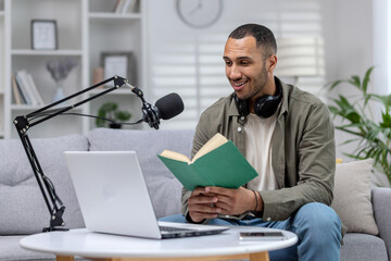 Young hispanic man in headphones sitting at home in front of a microphone and laptop and reading a...