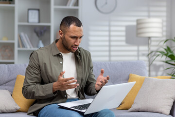 Worried young African-American man at home on sofa holding laptop and talking on video call with psychologist, undergoing online therapy and consultation