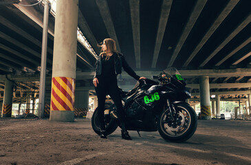 Woman in black leather jacket and jeans pants on outdoors parking with stylish sports motorcycle at sunset. Rider under highway