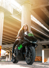 Fototapeta na wymiar Woman in black leather jacket and jeans pants on outdoors parking with stylish sports motorcycle at sunset. Rider under highway