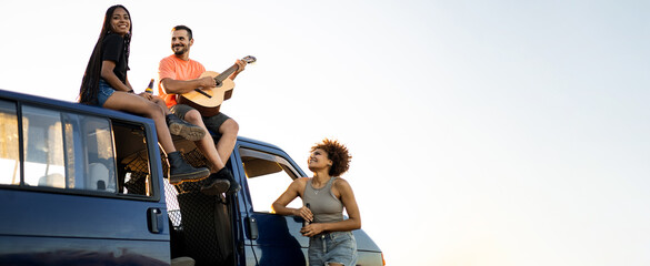 Advertising with copyspace to travel!A group of 3 young multi-ethnic people are having fun with a flamenco guitar while drinking beer happily near a camper van.Young people traveling with a camper van