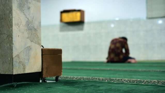 A blurred image of a man sitting in Jakarta's antique Faletehan mosque. He is praying alone on a green carpet not far from the pillar. Taken with a medium shot and handheld.