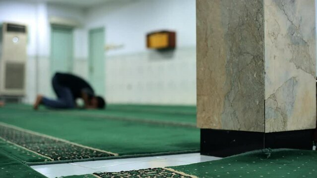A blurred image of a man praying at Jakarta's antique Faletehan mosque. He is praying alone on a green carpet not far from the pillar. Taken with a medium shot and handheld.