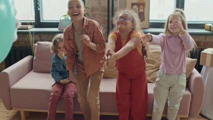 Medium full over-shoulder shot of group of 9-year-old multiethnic children sitting on couch at birthday party and watching unrecognizable entertainer in costume perform tricks with balloon