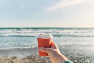 A young man holds a paper cup with coffee on the background of the sea. High-quality photo - Powered by Adobe