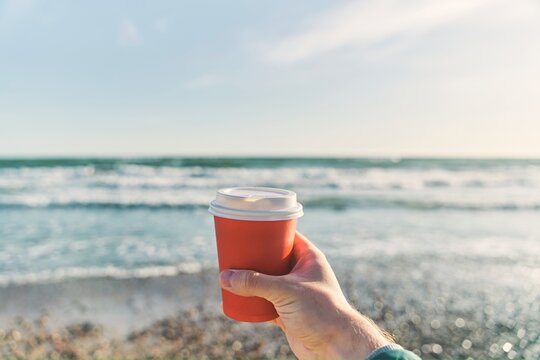 A young man holds a paper cup with coffee on the background of the sea. High-quality photo