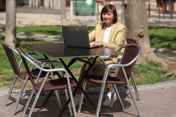 Elegant adult businesswoman works and type at laptop, making call on smartphone in an outdoor cafe at city. Concept of remote work communication from public place, digital freelance, modern lifestyle.
