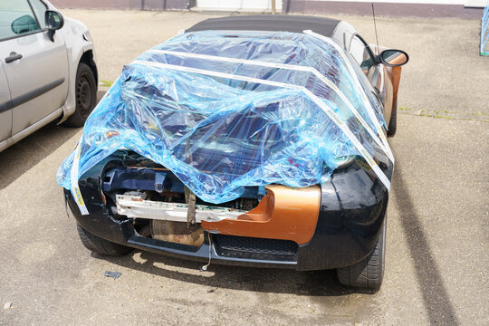 An accident-damaged car stands wrapped in film in a parking lot. Broken rear doors and bumper are visible.