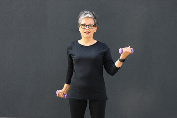 A mature woman in black sportswear is exercising with dumbbells in her hands.