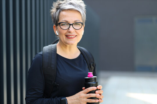 Portrait Of A Gray-haired Positive Woman In Glasses And Headphones With A Bottle Of Water After A Sports Workout.