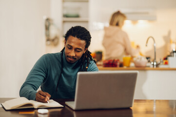 A dedicated interracial man is sitting at home and following an online lecture while his wife is cooking in a kitchen.