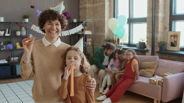 Medium portrait shot of happy curly Caucasian woman in casual jumper and birthday hat standing in living room hugging 8-year-old son, fooling around and blowing party horns while posing for photos