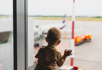 Toddler blond boy looks at the planes through the glass of a large window in the airport