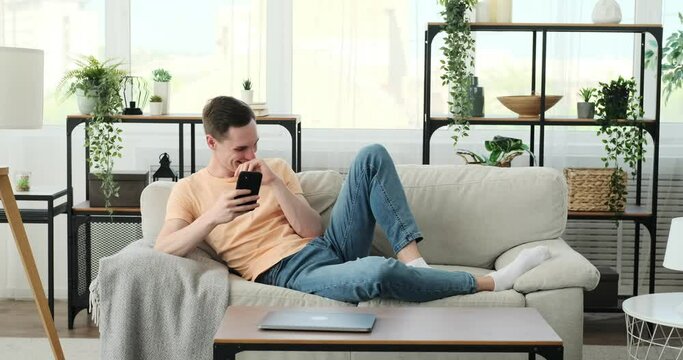 Caucasian man is seated comfortably on a couch, engrossed in his smartphone. With a relaxed posture, he effortlessly balances the device in his hand, his fingers tapping and scrolling with ease.