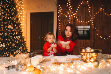 The concept of celebrating Christmas. Cheerful mom and little daughter in red pajamas cook Christmas cookies together and prepare for the holiday in the decorated kitchen of the house