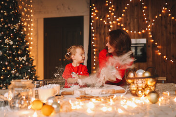 The concept of celebrating Christmas. Cheerful mom and little daughter in red pajamas cook Christmas cookies together and prepare for the holiday in the decorated kitchen of the house