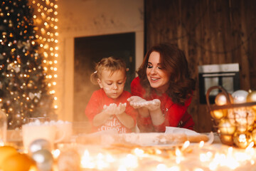 The concept of celebrating Christmas. Cheerful mom and little daughter in red pajamas cook Christmas cookies together and prepare for the holiday in the decorated kitchen of the house
