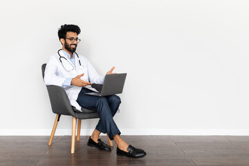 Handsome middle eastern young doctor sitting in armchair, using computer