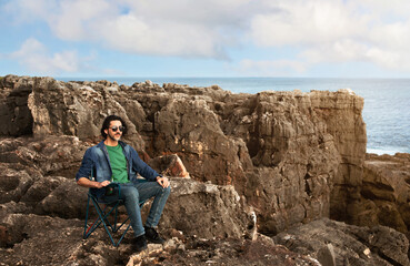 Naklejka na ściany i meble Handsome Young Man Relaxing In Camping Chair On Beachrocks Near Ocean