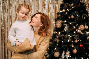 Fototapeta na wymiar A young mother holds a laughing daughter in her arms next to the Christmas tree.