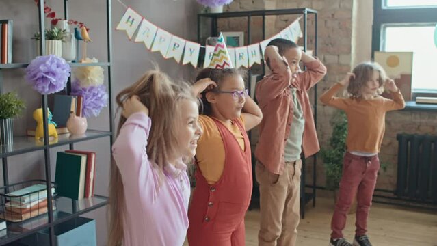 Medium shot of four young multiethnic boys and girls standing in line and performing fun physical exercises and tasks, while having fun at kids birthday party in family home