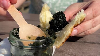 with a glass jar wooden spoon scoop black caviar on a bun on a croissant tasting tasting delicious food caviar fish. lumpfish with a wooden table close-up hands on the street picnic rich life