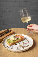 Glass of white wine in female hand and toast with salmon, avocado and poached egg