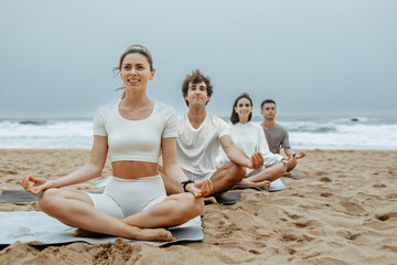 Happy women and men sitting in row in lotus padmasana pose with folded in mudra sign hands, meditating on the beach