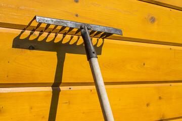 An old rusty rake is leaning against a yellow wooden wall and a contrasting shadow and copy space. ...