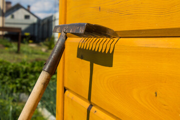 An old rusty metal rake near a yellow wooden wall and a bright contrasting shadow on a sunny day....