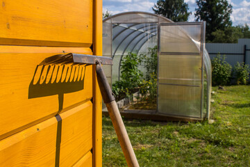 An old rusty metal rake near a yellow wooden wall and a bright contrasting shadow on a sunny day. The concept is gardening and agriculture