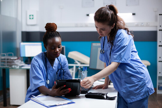 Diverse medical healthcare staff team looking at digital tablet screen in modern professional clinic office. African american nurse comparing data with coworker in hospital workplace