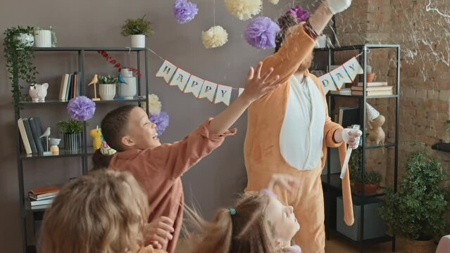 Medium slowmo shot of Caucasian man in cat character costume, with painted face entertaining four young children at house birthday party, making soap bubbles, kids laughing and catching them