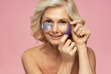 Caucasian blonde mature woman applying under eye patches on face looking at camera