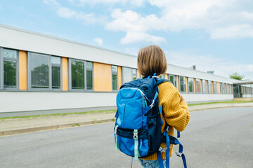 A view from the back of a girl with a school bag on her back is standing next to the school...