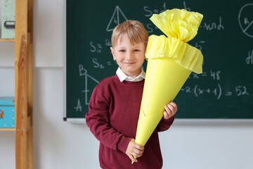 Happy little boy with yellow school cone in classroom near blackboard