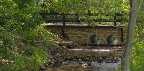 Mały most kamienny z drewnianymi poręczami wśród gęstej roślinności nad małą rzeczką ( Młynówką ) .